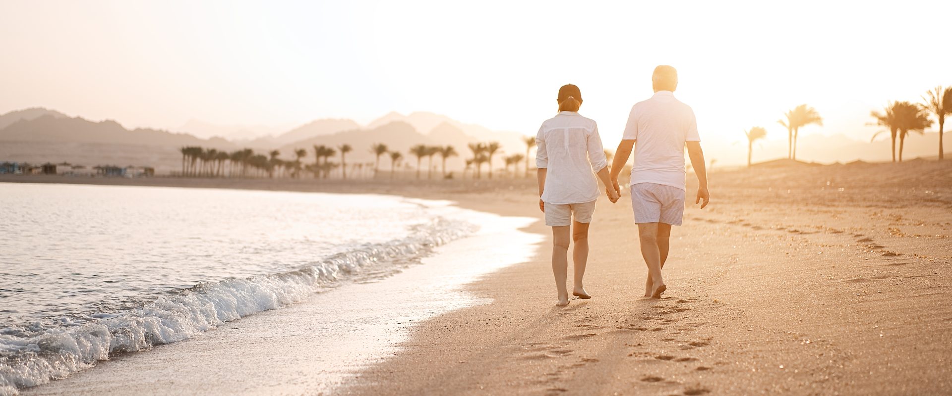 Happy mature senior couple walking and looking at each other on beach during sunset. Aging together and retirement lifestyle concept